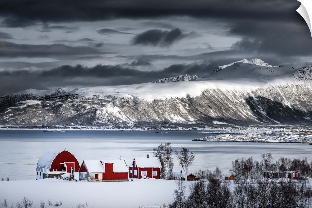 A photograph of a red building sitting on the shoreline of a snowy landscape with mountains in the distance.