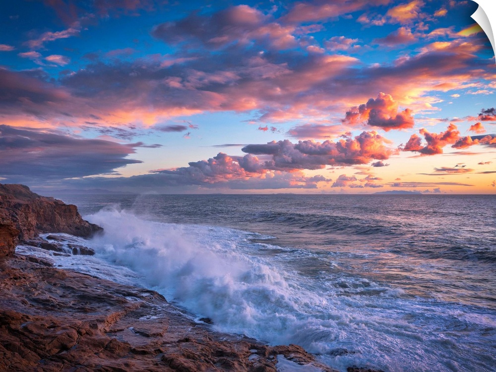 Beautiful photograph of a warm, cloudy sunset over the ocean with crashing waves onto the rocky shore.