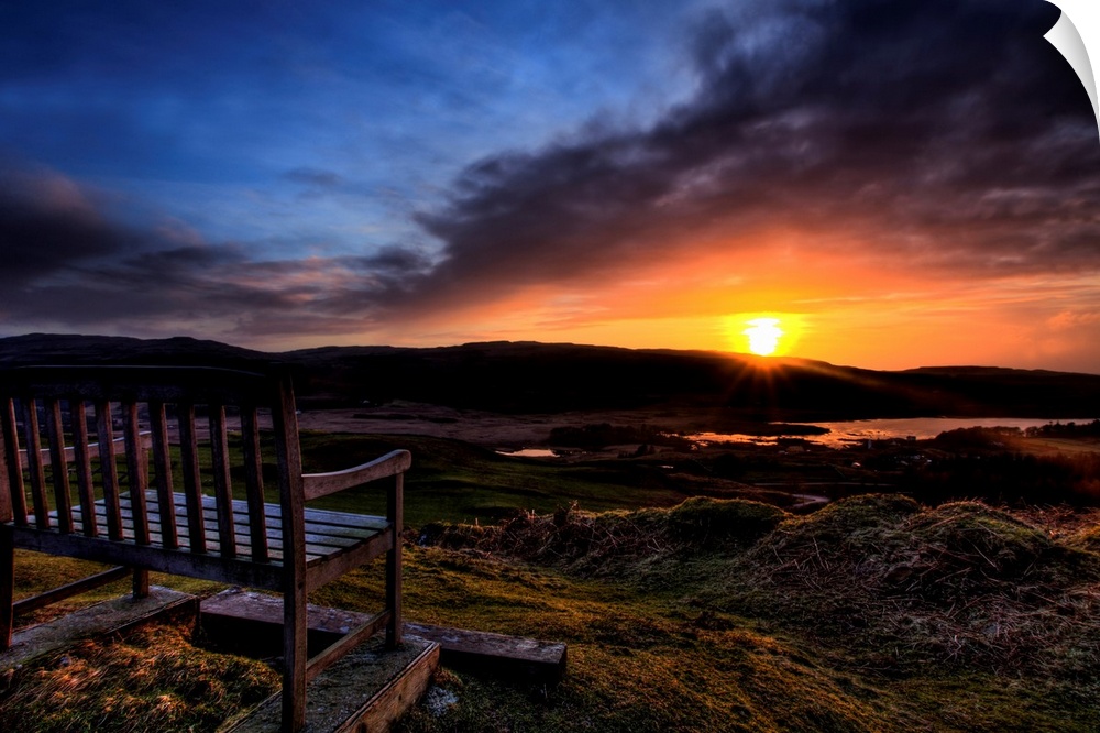A wooden seat looking out over a Scottish view to a Loch with a golden sunset and dramatic sky.