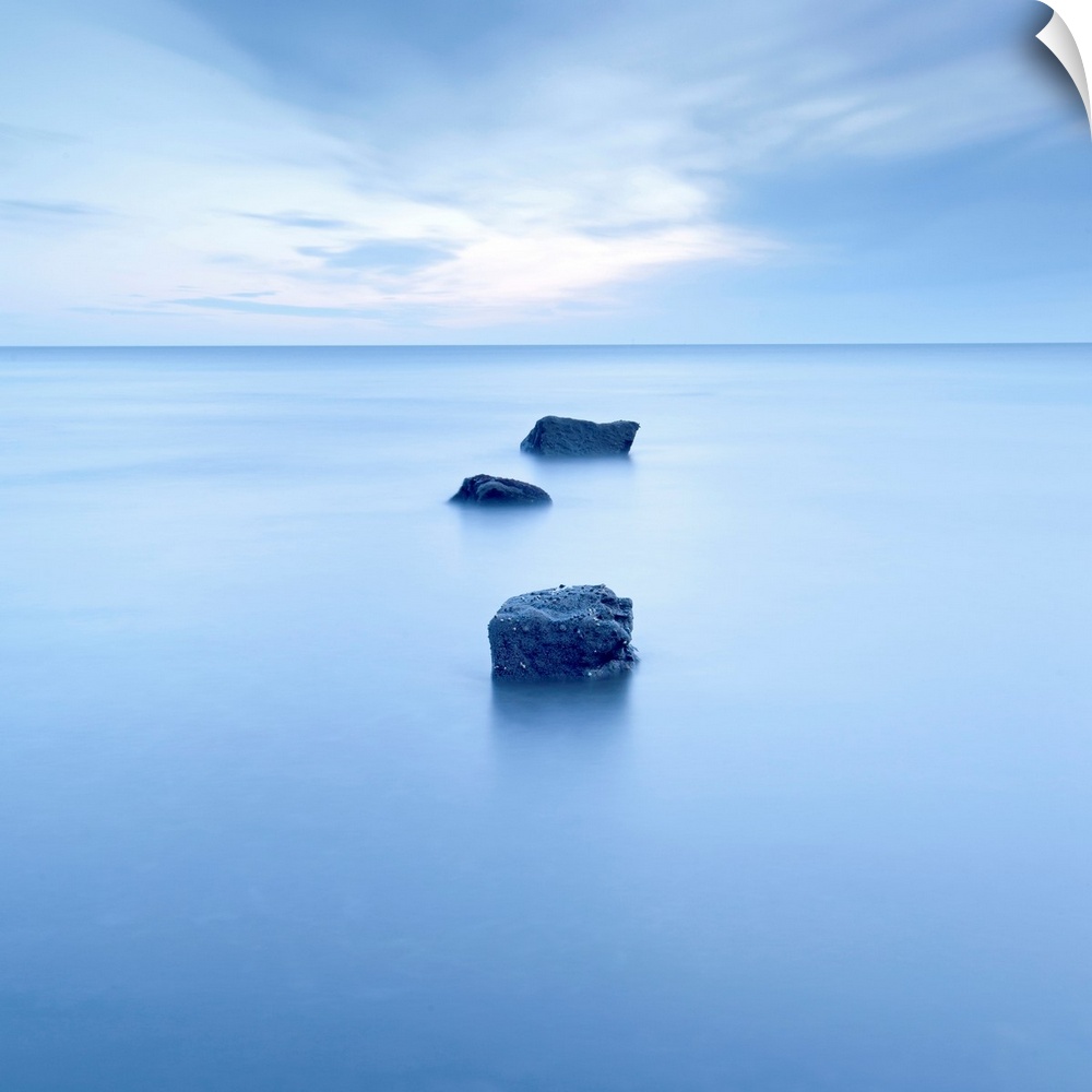 Large, square photograph of three rocks rising up from calm waters, beneath a partly cloudy sky.