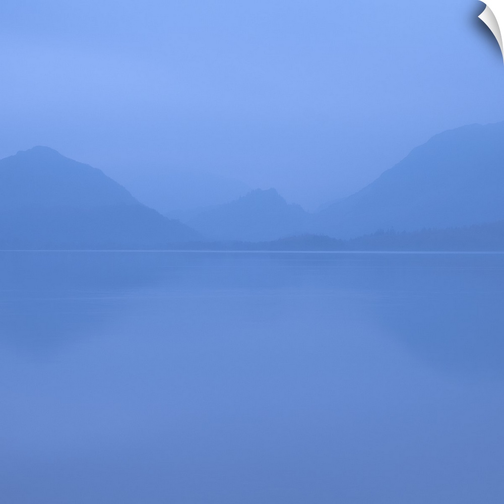 Square photograph of a blue foggy lake with mountains in the background.