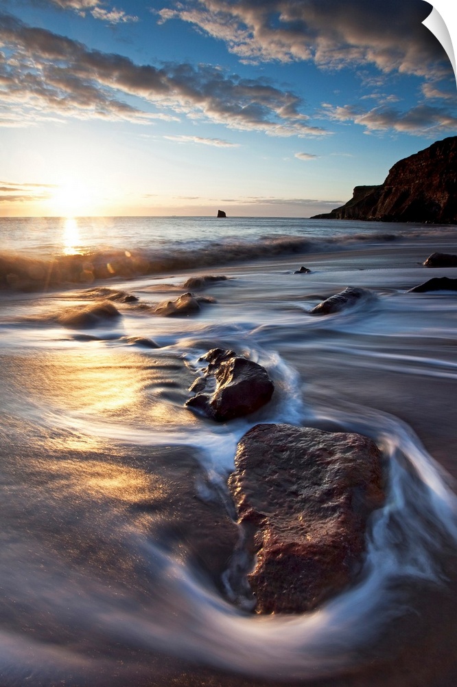 Vertical photograph on a large canvas of the sun rising over waters rushing onto a rocky beach, in Tyneside, Northern Engl...