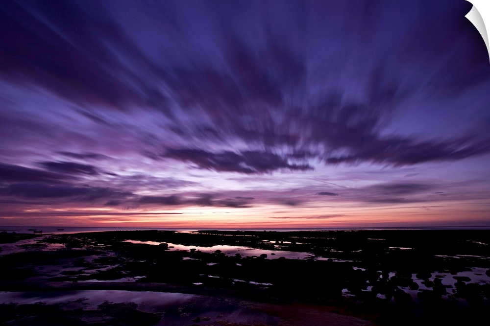 A dramatic seascape at dawn with deep purple magenta pink skies and clouds over wet sands.