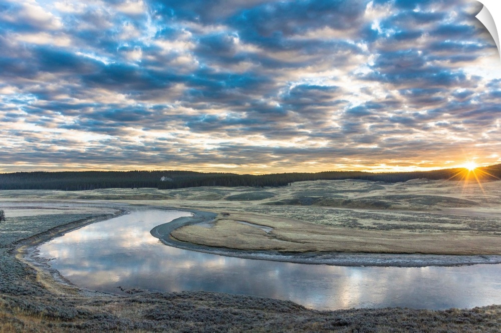 Fine art photograph of the sun on the horizon by the Yellowstone River with cloudy skies above.