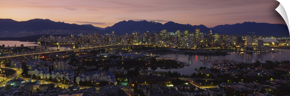 Panoramic photograph of skyline and waterfront at sunset with buildings lit up and mountain silhouettes in the distance.