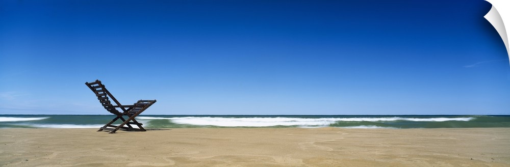 Huge panoramic piece of a lone beach chair skewed to the left sitting in the sand with waves crashing and a clear blue sky...