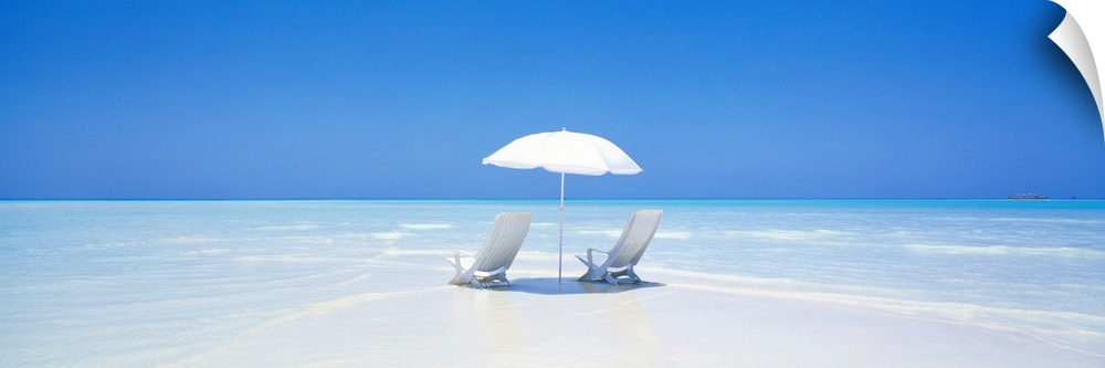 A panoramic photograph displaying two chairs sitting underneath an umbrella on a sandy beach in Maldives.  The clear water...