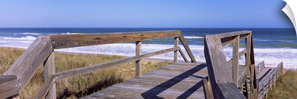 Panoramic picture taken of a small walking bridge headed down toward the beach with dunes on either side and the ocean str...