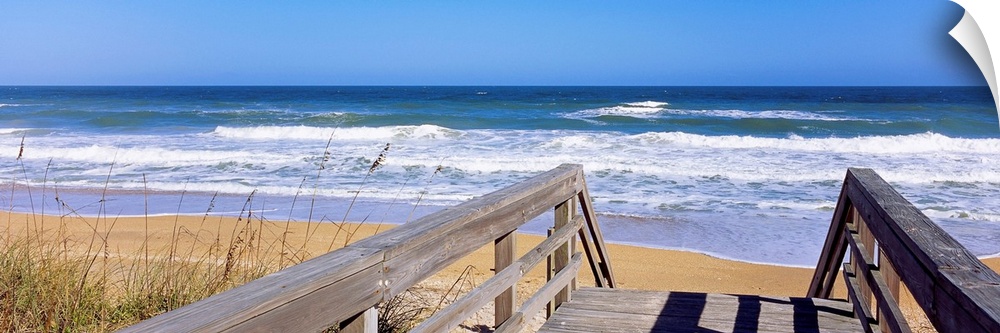 Panoramic photo of a wooden walkway leading to a beach shore with crashing waves during the day.
