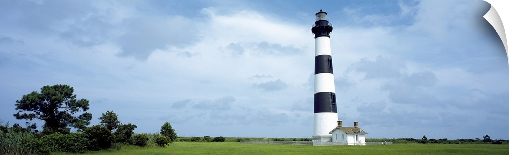 Panoramic photograph of striped Bodie Lighthouse surrounded by a green landscape, beneath a cloudy blue grey sky.