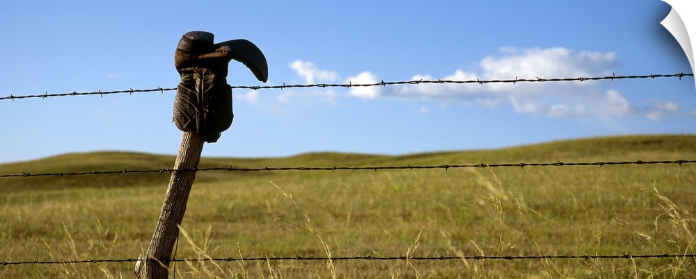 Panoramic photograph of cowboy boot hanging upside down on a cable fence with grass covered hills and a cloudy sky in the ...