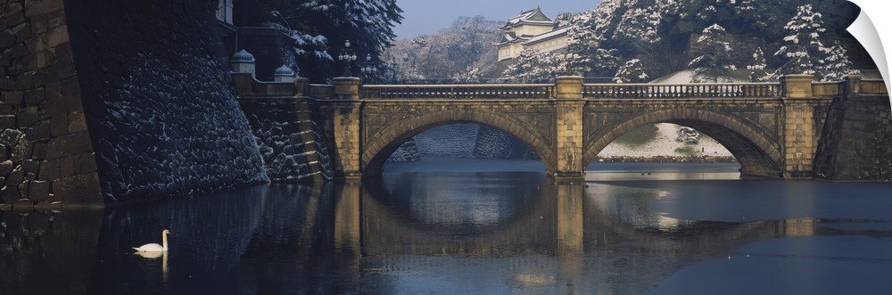 Panoramic photograph of the stone Bridge Nijubashi over calm waters, surrounded by a snow covered landscape as a swan floa...