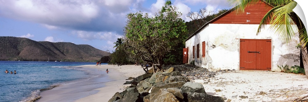 Wide angle photograph taken of a building sitting on a beach with rocks sitting just in front of it and the ocean with peo...