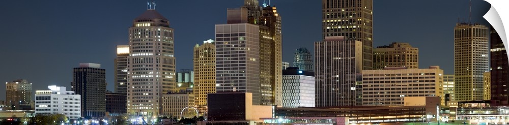 Oversize horizontal photograph of lit skyscrapers against a deep blue sky at night, in Detroit, Michigan.