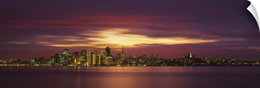 Giant, panoramic photograph from a distance of the San Francisco skyline, lit up beneath a colorful sunset.