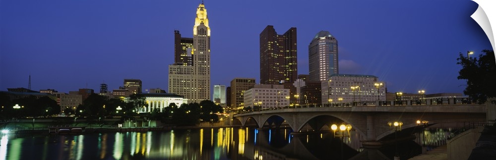 The city of Columbus Ohio is illuminated under a night sky and photographed in panoramic view from across a body of water.