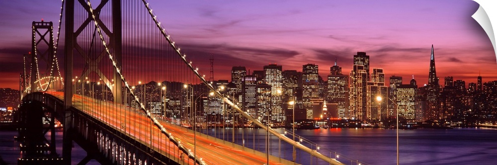 Panoramic photograph of the San Francisco Bay Bridge and the brightly lit city skyline at dusk.