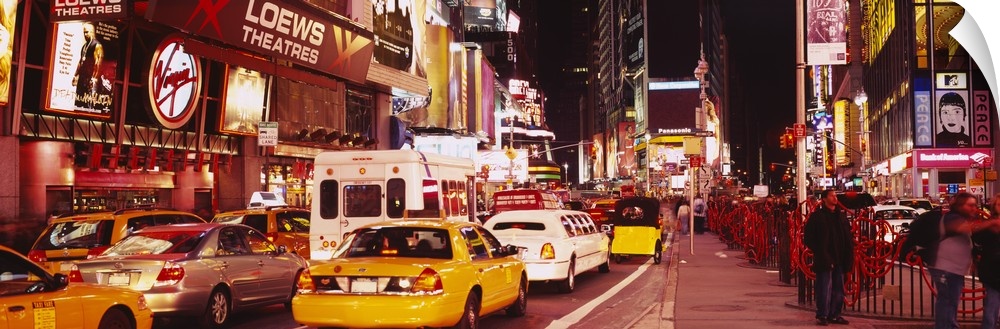 Panoramic photograph of many cars and taxis filling the street in Times Square, surrounded by the bright lights and billbo...