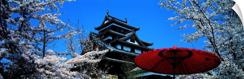 Beautiful trees are pictured surrounding a Japanese castle with a red umbrella near the bottom right hand side of the phot...