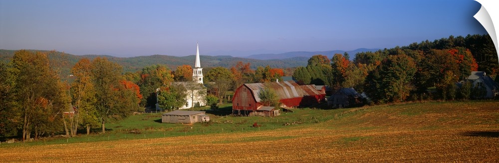 This panoramic photograph is of a barn and a church surrounded by autumn trees with large hills in the distance.