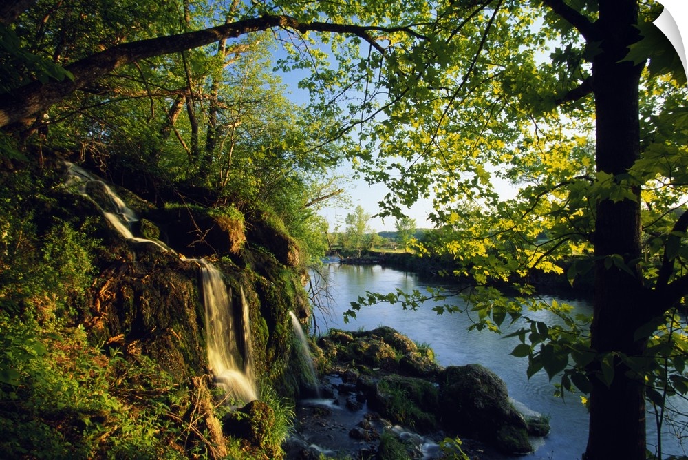 Small waterfall with mossy rocks and leafy trees on the edge of a river in the early morning.