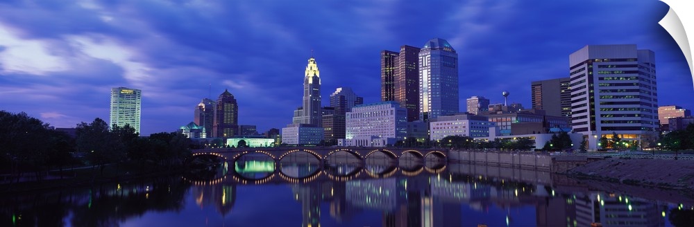 Panoramic photo of downtown Columbus, Ohio with the tall buildings and lit  up bridge reflecting in the river.