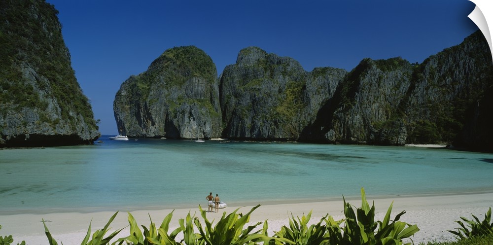 A breathtaking photograph of a beach in Thailand surrounded by mountains with a couple at the edge of the clear water.