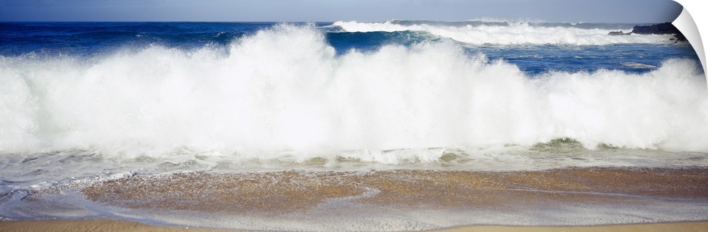 Panoramic photograph of large waves crashing into the beach, bright blue water in the background, in Waimea Bay, Hawaii.