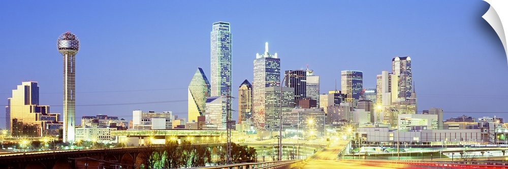 Giant, landscape photograph of a highway road leading toward the lit up Dallas skyline against a blue sky, at dusk.