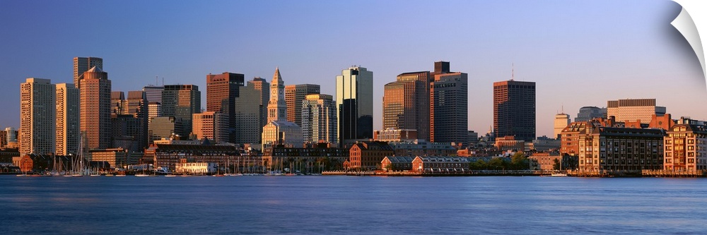 Large panoramic photograph of the Boston skyline from across the river with the sun hitting the buildings as it rises.