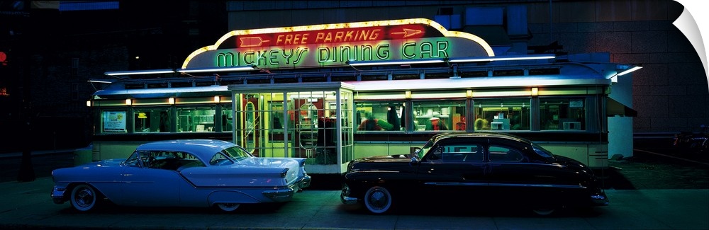 Panoramic photograph of two vintage cars sitting outside of a lit up eatery at night with neon signs.