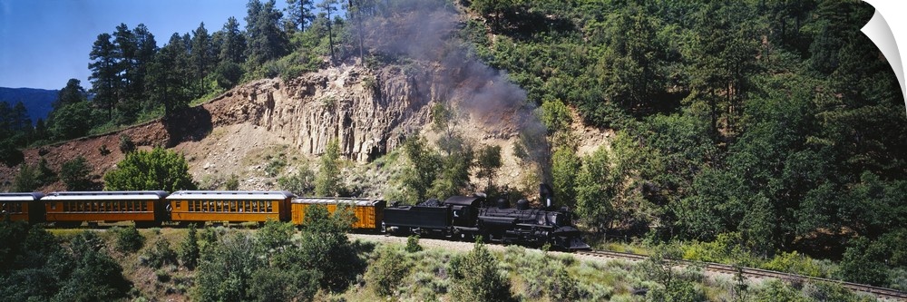 This is a panoramic photograph of a vintage engine and train cars passing through the mountains on a sunny day.