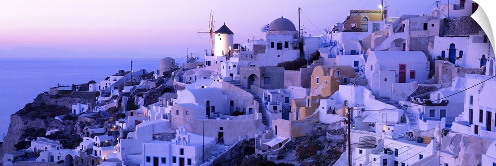 Wide angle photograph of various buildings on a sloping hillside near the coast, in Santorini, Greece, at sunset.