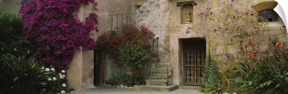 Panoramic image of an Italian style stone house with large flowering bushes crawling up the side and a wrought iron stairc...