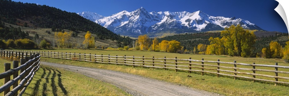 Panoramic photograph of dirt path lined by a wooden gate with snow covered mountains in the distance under a clear sky.