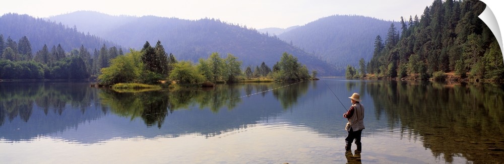 A fisherman stands in shallow water with his line out. There is a beautiful view of trees and large hills in the distance.