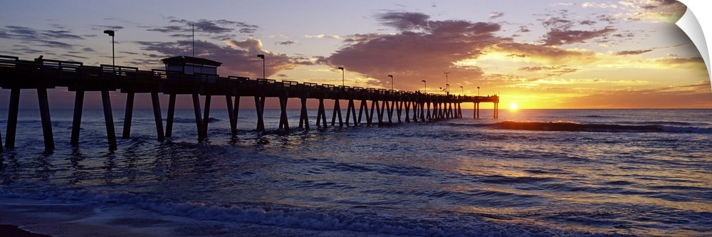 A pier that reaches far out into the ocean is photographed from the beach with the sun just setting on the horizon.