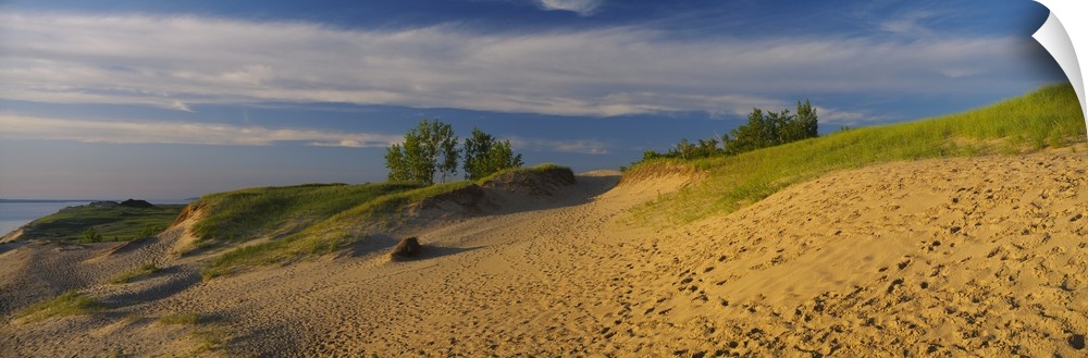 Sloping sand dunes covered in beach grass and footprints under wispy clouds at sunset at the edge of a lake.