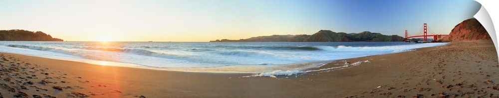 Long panoramic photo of a beach with the golden gate bride on the far right and the setting sun on the far left.