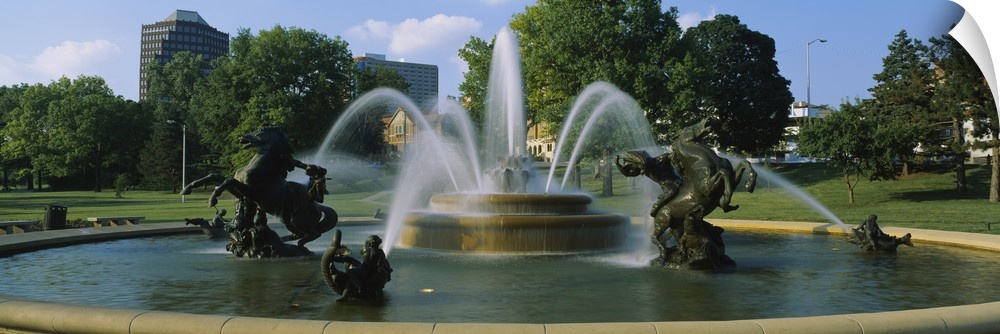 A large fountain with various statues in it is photographed in panoramic view with a park shown in the background.