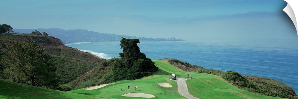 This wall hanging is a panoramic photograph of a golf green overlooking the ocean from the top of a hill.