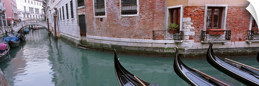The front of gondola boats are pictured in the water with buildings to either side of the canal where more boats are shown.