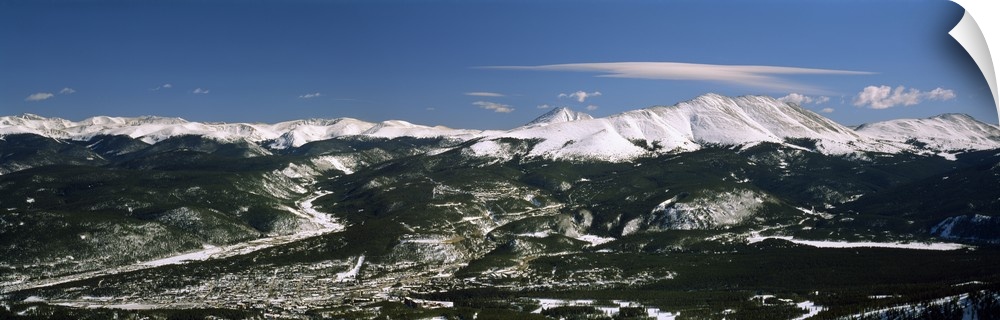 This wide angle photograph is an aerial shot of a snow covered mountain range with vast land and a town in front of them.