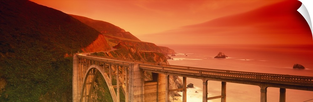 Oversized, horizontal photograph of the Bixby Bridge, next to the cliffs of Big Sur, California, a fiery sunset casts a wa...