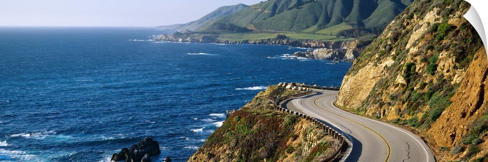 Big, panoramic landscape photograph of curving Route 1 on a hillside, along the coast of Big Sur, California.