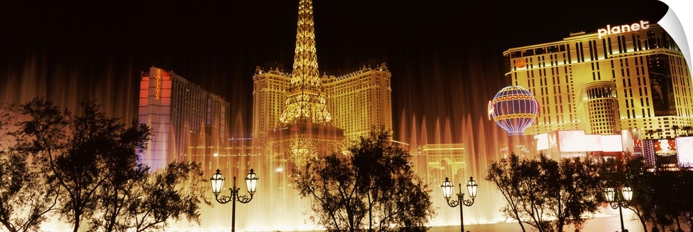 Panoramic photograph of ""Sin City"" skyline and Bellagio fountains with tree and street lamp silhouettes in the foreground.
