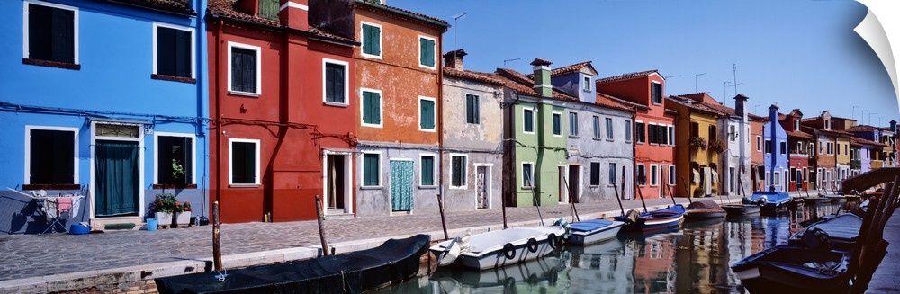Panoramic photograph of colorful houses lined up next to each other on a lagoon with boats tied up in the water.