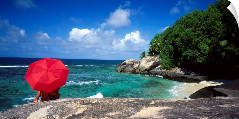 A woman holding a red umbrella sits on a large rock and looks out over the ocean with the coast just to her right.