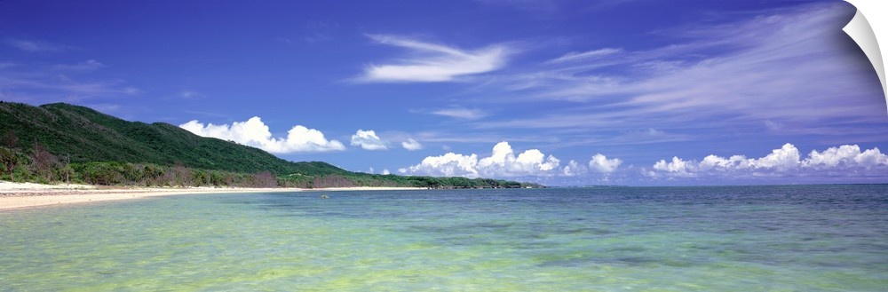 Panoramic photograph of ocean with shoreline and grass covered mountains in the distance under a cloudy sky.