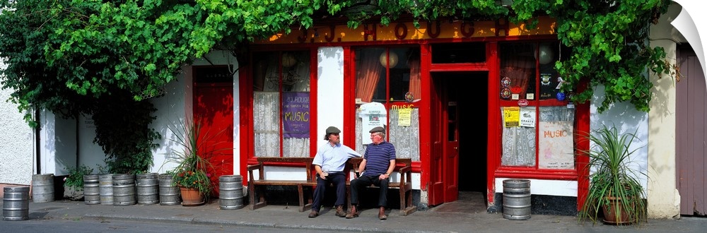 Panoramic picture snapped of two men as they sit on a bench outside a bar in Ireland. The front of the bar is lined with b...